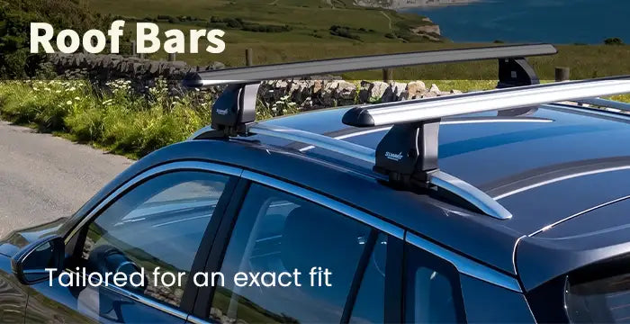A black & silver roof bar system installed on the roof of a dark blue car, shown outdoors on a road with green hills and water in the background. Text overlays read 'Roof Bars' and 'Tailored for an exact fit'.
